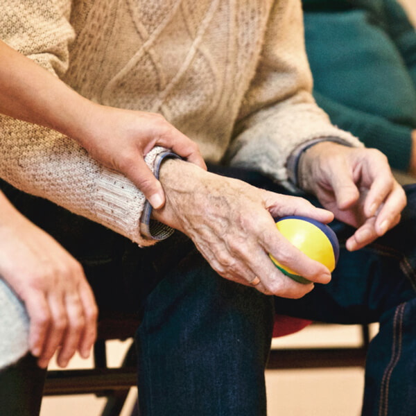 Ältere Hand drückt Therapiekugel, Therapeut stützt Handgelenk