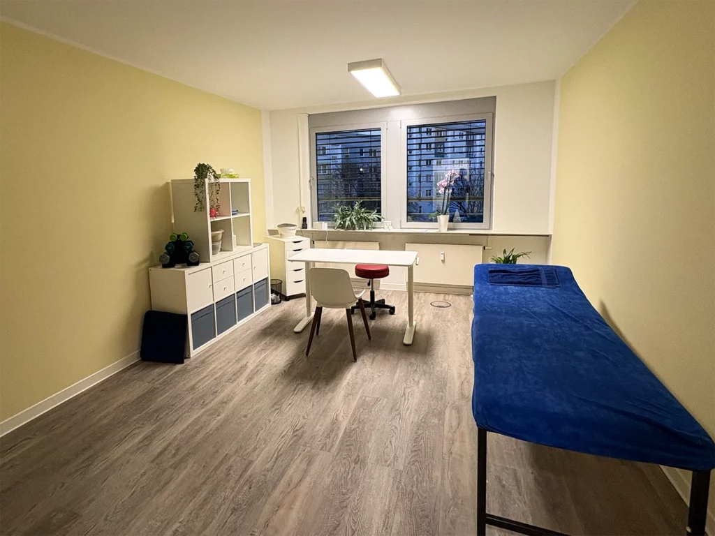 Heller Therapieraum mit Behandlungsliege, Schreibtisch, Stühlen und Regal, ausgestattet mit Pflanzen und Therapiematerialien.