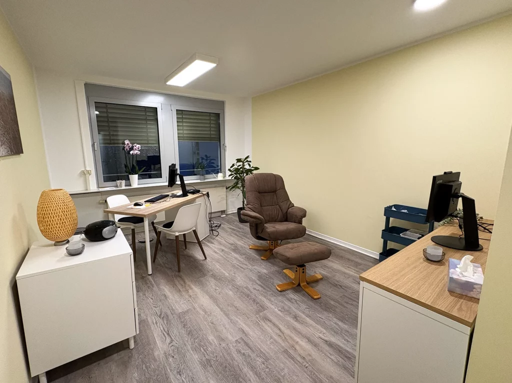 Modern eingerichtetes Behandlungszimmer mit Schreibtisch, Monitor, Relaxsessel und dekorativen Elementen vor einem Fenster