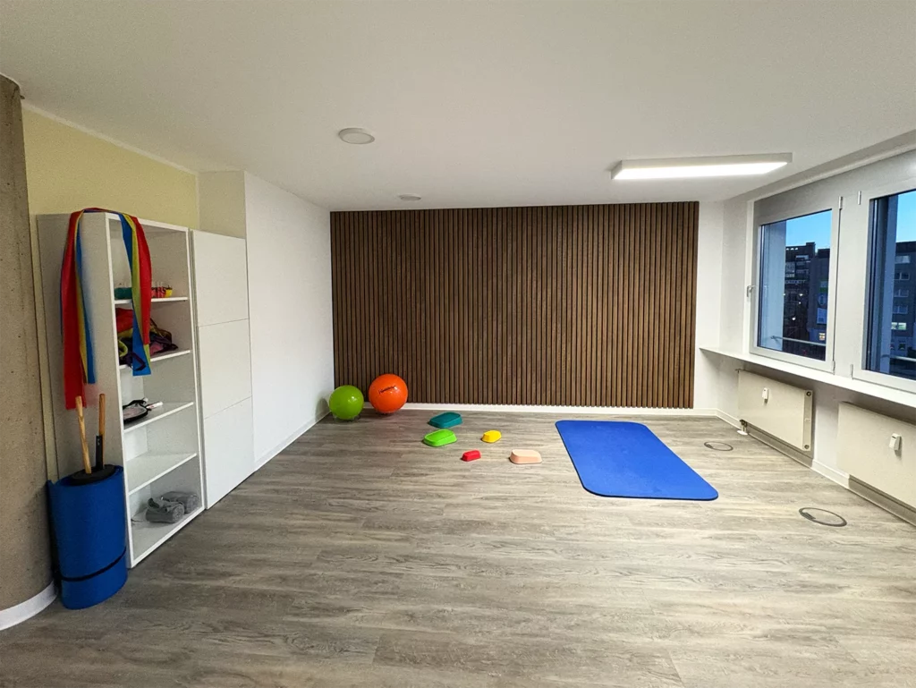 Bewegungsraum mit Gymnastikmatte, Therapieball, Spielgeräten und großem Fenster, geeignet für körperliche Aktivitäten.